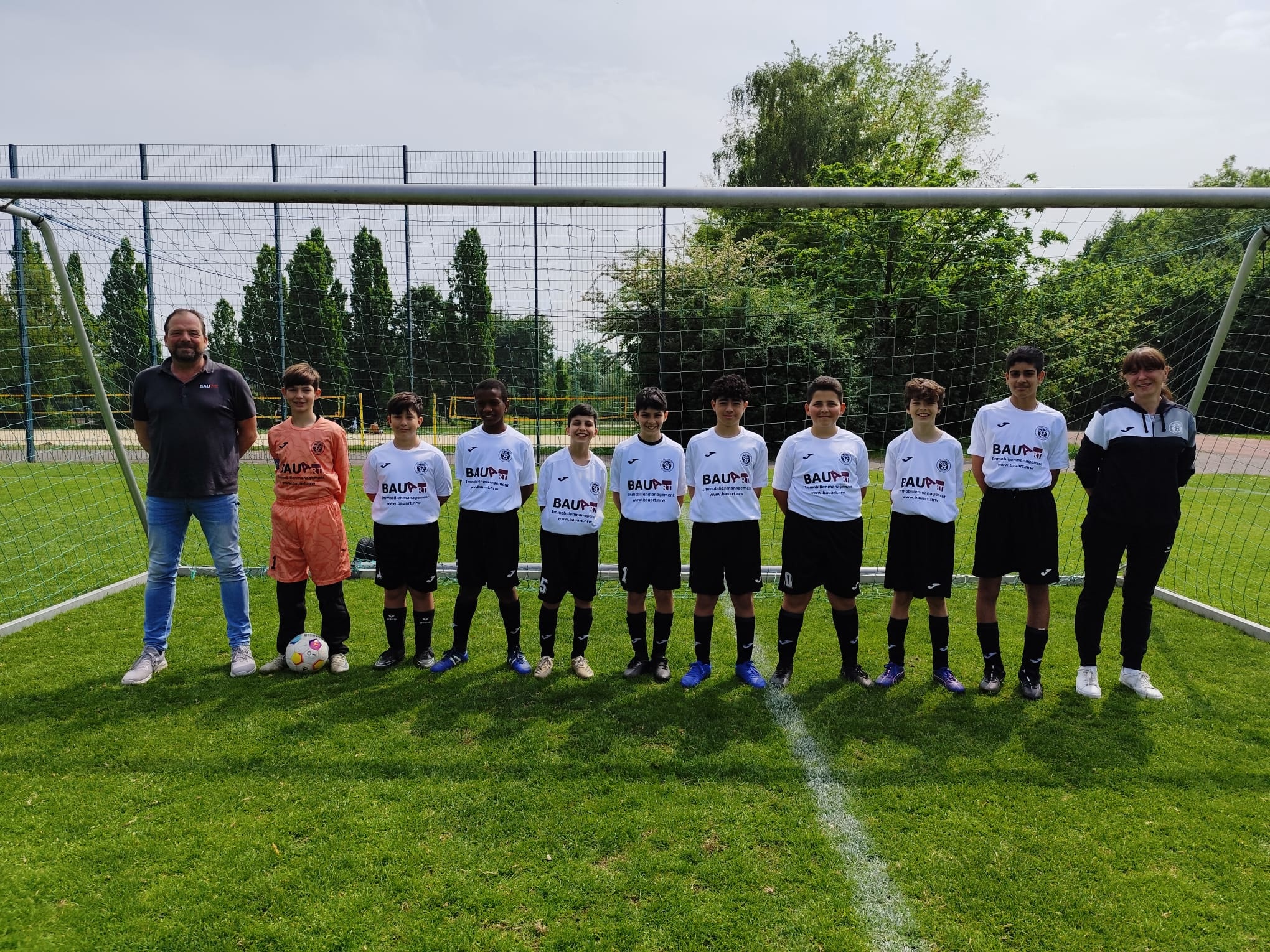Teamfoto mit dem neuen Sponsor BauArt. Acht Kinder stehen in weißen Trikots mit schwarzen Hosen in einem Tor. Links steht der männliche Sponsor. Rechts die Trainerin. Der Torhüter hat ein orangenes Trikot an. Er ist der zweite Spieler von links auf dem Foto.